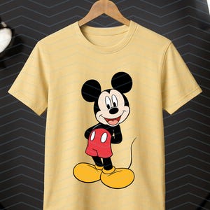May include: A light yellow short-sleeve t-shirt featuring a centered graphic of Mickey Mouse. The character is depicted with his signature black ears, red shorts with two white buttons, and large yellow shoes, standing against a plain background.