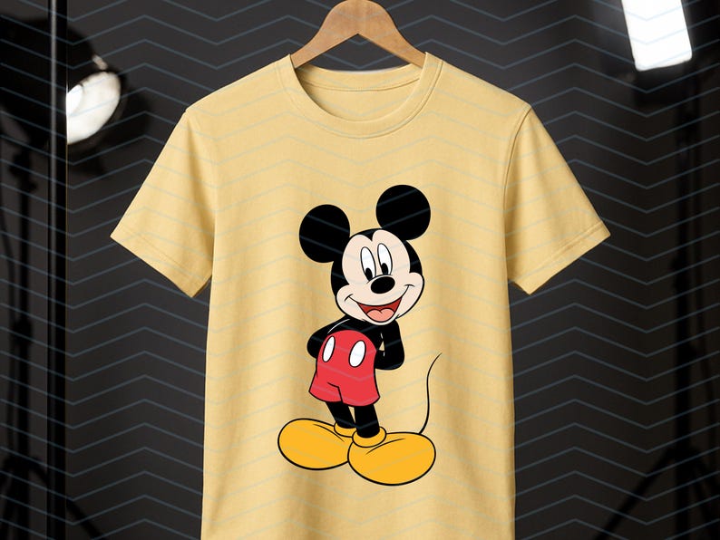May include: A light yellow short-sleeve t-shirt featuring a centered graphic of Mickey Mouse. The character is depicted with his signature black ears, red shorts with two white buttons, and large yellow shoes, standing against a plain background.