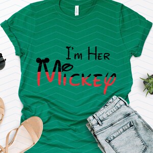 Op de afbeelding: Een groen T-shirt met de tekst "I'm Her Mickey" in zwarte en rode letters. Het shirt ligt plat op een witte achtergrond met een paar sandalen, een zonnebril en een spijkerbroek.