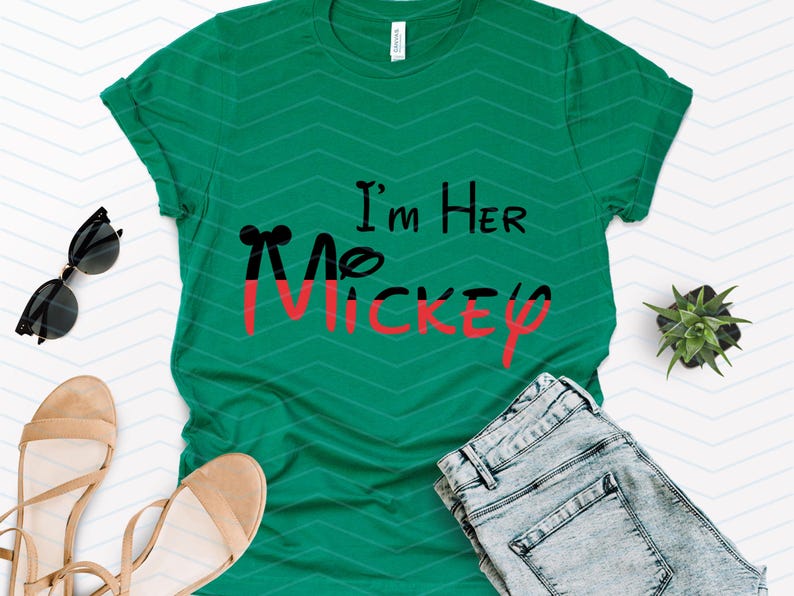 Op de afbeelding: Een groen T-shirt met de tekst "I'm Her Mickey" in zwarte en rode letters. Het shirt ligt plat op een witte achtergrond met een paar sandalen, een zonnebril en een spijkerbroek.