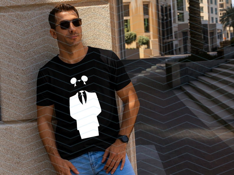 May include: A black short-sleeved t-shirt featuring a white graphic print of a stylized suit jacket, a necktie, and a pair of circular ears positioned above the collar. The design is centered on the chest of the garment.