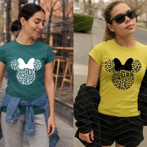 May include: Two women wearing t-shirts with a Minnie Mouse design. One shirt is teal with a white leopard print design, and the other is yellow with a black leopard print design. Both shirts have a bow.
