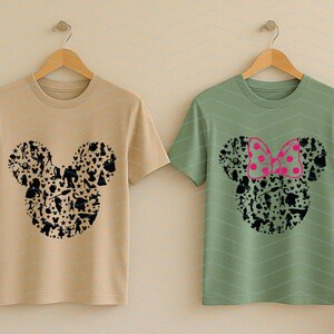 May include: Two short-sleeve crew neck t-shirts hanging on wooden hangers. The left shirt is beige with a black graphic of a Mickey Mouse head silhouette filled with various character shapes. The right shirt is sage green with a similar black silhouette graphic featuring a bright pink polka-dot bow.