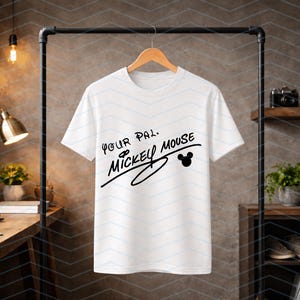May include: A white short-sleeved t-shirt hanging on a wooden hanger. The front of the shirt features black text that reads Your Pal, Mickey Mouse, accompanied by a small black silhouette of the iconic Mickey Mouse ears icon.