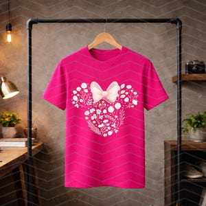 May include: A bright pink t-shirt featuring a floral Minnie Mouse design. The design includes a white bow and floral elements arranged in the shape of Minnie Mouse's head. The shirt is hanging on a wooden hanger.