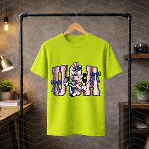May include: A lime green short-sleeve t-shirt featuring a graphic of Mickey Mouse wearing a patriotic hat. The letters USA are printed in a red and white striped pattern with blue stars, accented by two blue patterned bows on the sides.