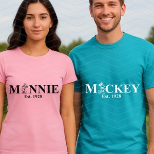 Pu&ograve; includere: Maglietta rosa con il nome "MINNIE" in nero, un'immagine di un cartone animato e "Est. 1928" stampato sotto. Maglietta verde acqua con il nome "MICKEY" in bianco, un'immagine di un cartone animato e "Est. 1928" stampato sotto.