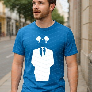 May include: A bright blue short-sleeved t-shirt featuring a large white graphic print on the chest. The graphic depicts a stylized suit jacket with a necktie and a pair of circular mouse-like ears positioned above the collar area.