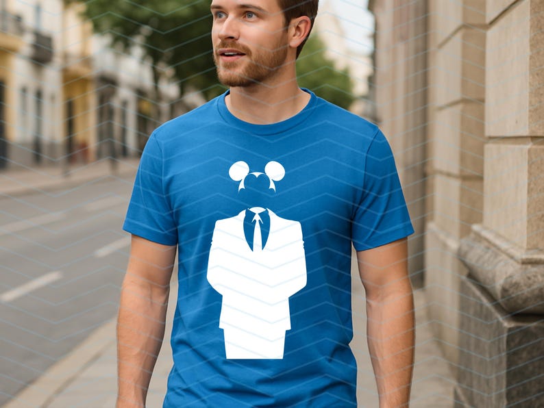 May include: A bright blue short-sleeved t-shirt featuring a large white graphic print on the chest. The graphic depicts a stylized suit jacket with a necktie and a pair of circular mouse-like ears positioned above the collar area.