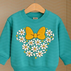 Puede incluir: Una sudadera turquesa con un dise&ntilde;o de lazo y margaritas. El dise&ntilde;o presenta un lazo amarillo y margaritas blancas dispuestas en forma de cabeza de Mickey Mouse. La sudadera tiene mangas largas y cuello redondo.