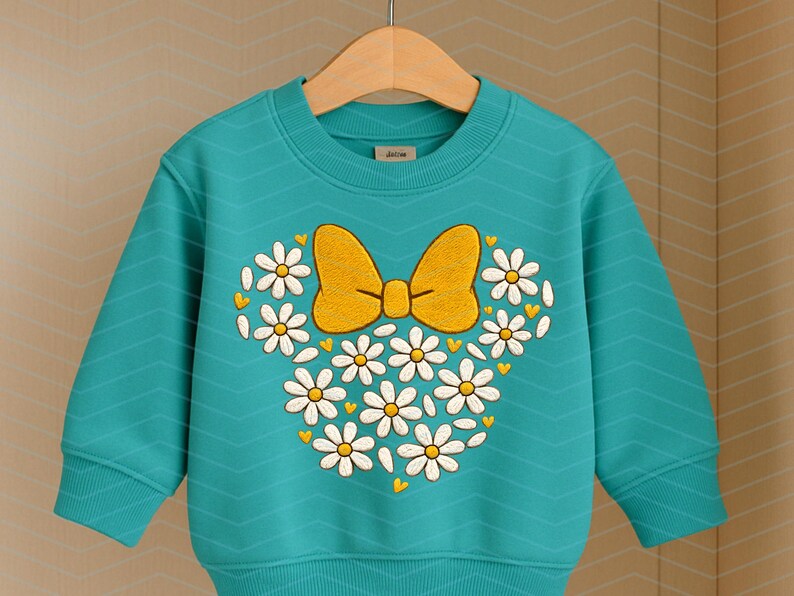 Puede incluir: Una sudadera turquesa con un dise&ntilde;o de lazo y margaritas. El dise&ntilde;o presenta un lazo amarillo y margaritas blancas dispuestas en forma de cabeza de Mickey Mouse. La sudadera tiene mangas largas y cuello redondo.