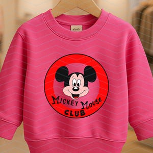 May include: A bright pink long-sleeve sweatshirt featuring a circular red graphic on the chest. The graphic displays the face of Mickey Mouse and the black text "MICKEY MOUSE CLUB". The garment has a ribbed crew neckline, cuffs, and hem.