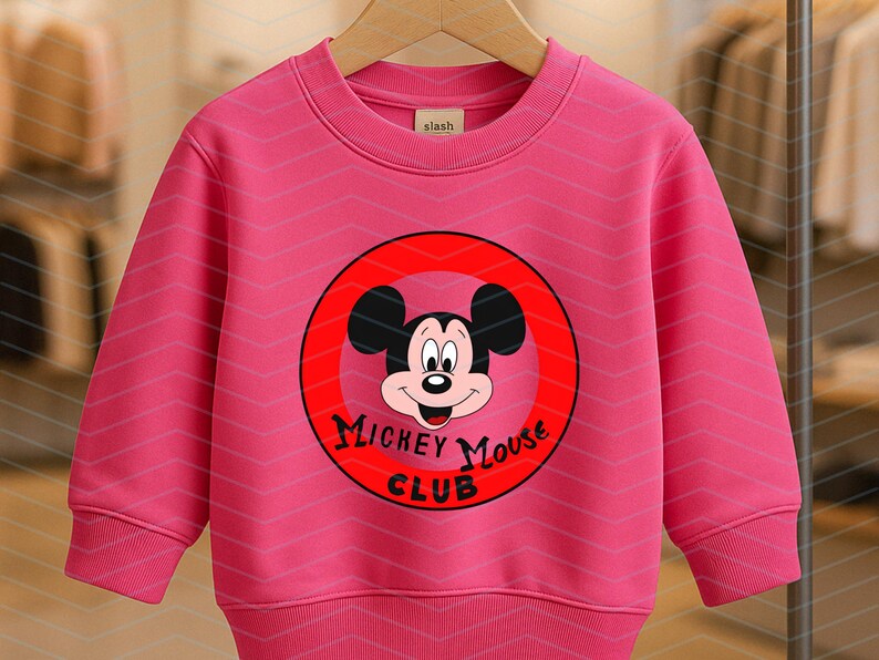 May include: A bright pink long-sleeve sweatshirt featuring a circular red graphic on the chest. The graphic displays the face of Mickey Mouse and the black text "MICKEY MOUSE CLUB". The garment has a ribbed crew neckline, cuffs, and hem.