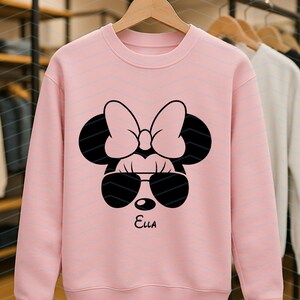 May include: A light pink sweatshirt featuring a black graphic of a cartoon mouse face with sunglasses and a bow. The name "Ella" is printed in black script below the graphic. Other sweatshirts are visible in the background.