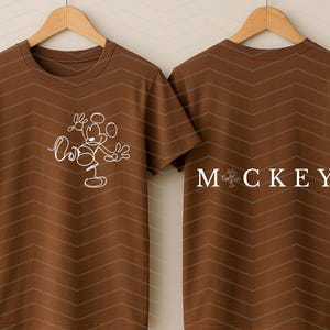 Puede incluir: Una camiseta marr&oacute;n de manga corta con un gr&aacute;fico de l&iacute;neas blancas de un rat&oacute;n de dibujos animados en la parte delantera. La parte trasera muestra el texto "MOCKEY" en letras may&uacute;sculas blancas, con un icono de rat&oacute;n sustituyendo la letra O.