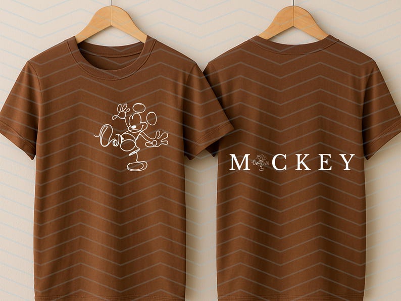 Puede incluir: Una camiseta marr&oacute;n de manga corta con un gr&aacute;fico de l&iacute;neas blancas de un rat&oacute;n de dibujos animados en la parte delantera. La parte trasera muestra el texto "MOCKEY" en letras may&uacute;sculas blancas, con un icono de rat&oacute;n sustituyendo la letra O.