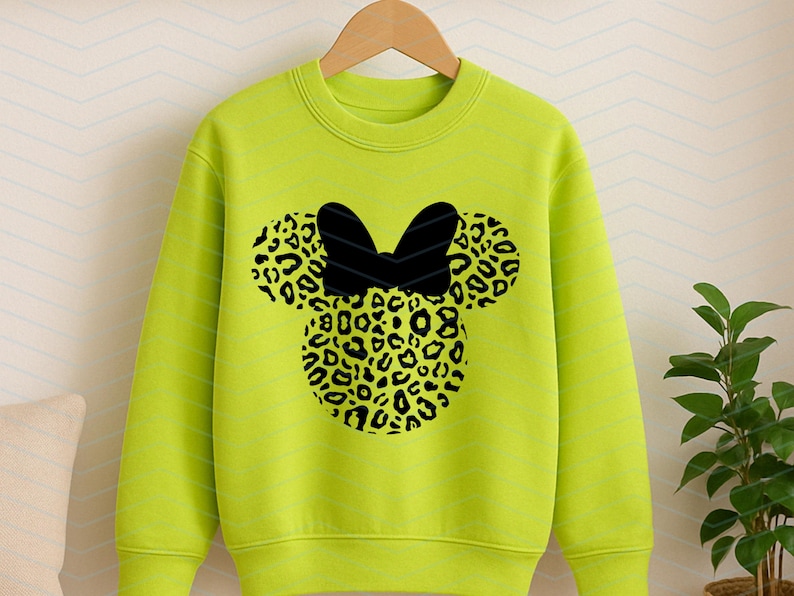 May include: A neon green sweatshirt featuring a black bow and a leopard print design in the shape of a mouse head. The sweatshirt is hanging on a wooden hanger.