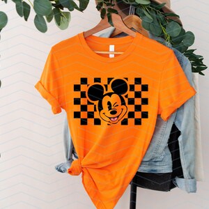 May include: Orange t-shirt with a black and white checkered pattern and a cartoon image of Mickey Mouse with a mischievous grin.