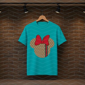 Puede incluir: Una camiseta de manga corta color verde azulado con un gr&aacute;fico central de una silueta de cabeza de rat&oacute;n con un lazo rojo. La silueta tiene un patr&oacute;n repetitivo en color tostado y marr&oacute;n con una franja vertical verde y roja a la derecha.