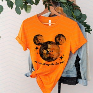 Puede incluir: Camiseta naranja con una silueta negra de Mickey Mouse hecha de bolas de discoteca. El texto "Me and Mickey vibe like that" est&aacute; impreso debajo de la silueta.