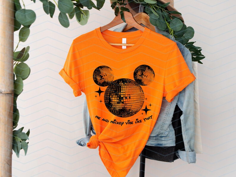 Puede incluir: Camiseta naranja con una silueta negra de Mickey Mouse hecha de bolas de discoteca. El texto "Me and Mickey vibe like that" est&aacute; impreso debajo de la silueta.
