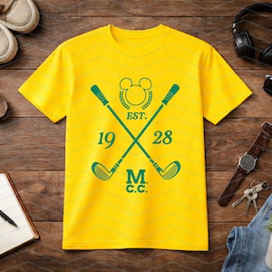 May include: Yellow t-shirt with a green golf-themed design. The design includes crossed golf clubs, the text "EST. 1928 M.C.C.", and a Mickey Mouse head silhouette. The shirt is displayed on a wooden surface with accessories.