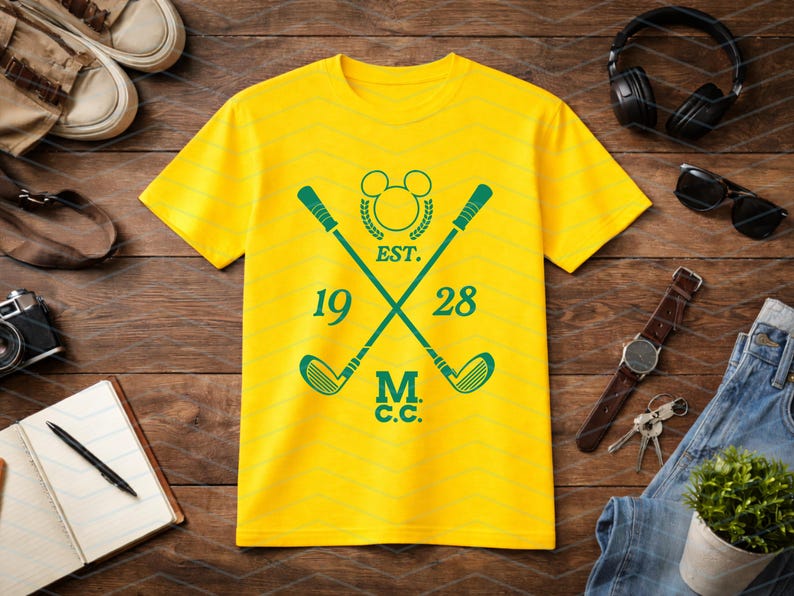 May include: Yellow t-shirt with a green golf-themed design. The design includes crossed golf clubs, the text "EST. 1928 M.C.C.", and a Mickey Mouse head silhouette. The shirt is displayed on a wooden surface with accessories.