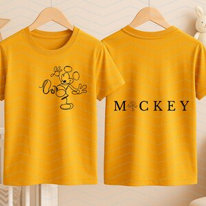 Puede incluir: Una camiseta de manga corta color amarillo mostaza mostrada de frente y de espaldas. El frente presenta un dibujo de l&iacute;nea negra de Mickey Mouse. La parte posterior muestra el texto negro "M CKEY" con un peque&ntilde;o gr&aacute;fico reemplazando la letra I.