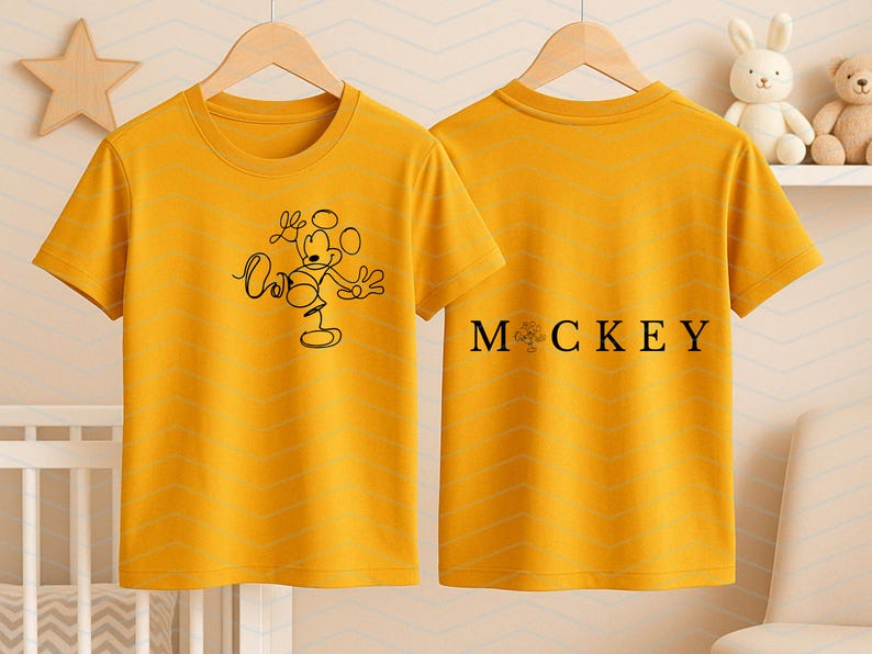 Puede incluir: Una camiseta de manga corta color amarillo mostaza mostrada de frente y de espaldas. El frente presenta un dibujo de l&iacute;nea negra de Mickey Mouse. La parte posterior muestra el texto negro "M CKEY" con un peque&ntilde;o gr&aacute;fico reemplazando la letra I.