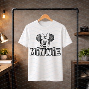 May include: White t-shirt with a black and white Minnie Mouse design. The graphic shows Minnie's face, bow, and the name "MINNIE". The t-shirt is displayed on a wooden hanger.