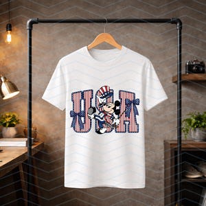 May include: A white short-sleeved t-shirt featuring a graphic of a cartoon mouse wearing a patriotic top hat. The letters USA are printed in a red and white striped pattern with blue star accents and blue bows on the sides of the letters.