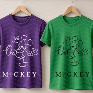 Puede incluir: Dos camisetas de manga corta y cuello redondo colgadas en perchas de madera. La camiseta de la izquierda es morada con un dibujo de l&iacute;nea continua blanca de un rat&oacute;n de dibujos animados y el texto MOCKEY. La de la derecha es verde con negro.