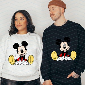 May include: Two white sweatshirts with a black and red Mickey Mouse graphic on the front. The sweatshirts are shown on two people.