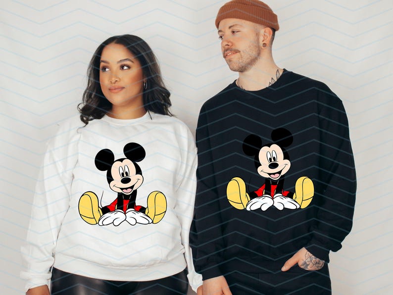 May include: Two white sweatshirts with a black and red Mickey Mouse graphic on the front. The sweatshirts are shown on two people.