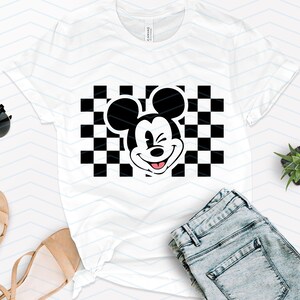 May include: White t-shirt with a black and white checkered pattern and a black and white image of Mickey Mouse winking. Mickey Mouse is wearing a red tongue.