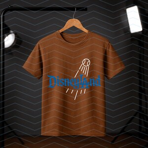 May include: A brown t-shirt featuring the word "Disneyland" in blue script, and a baseball graphic with lines. The shirt is suspended on a wooden hanger against a dark backdrop.