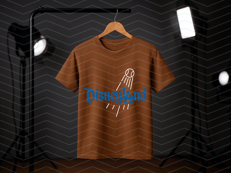 May include: A brown t-shirt featuring the word "Disneyland" in blue script, and a baseball graphic with lines. The shirt is suspended on a wooden hanger against a dark backdrop.