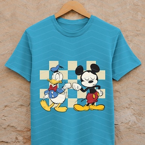May include: A turquoise t-shirt featuring a cartoon illustration of Donald Duck and Mickey Mouse bumping fists. Donald Duck is wearing a blue sailor suit and Mickey Mouse is in red shorts. The background has a checkered pattern.