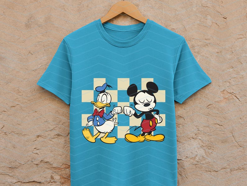 May include: A turquoise t-shirt featuring a cartoon illustration of Donald Duck and Mickey Mouse bumping fists. Donald Duck is wearing a blue sailor suit and Mickey Mouse is in red shorts. The background has a checkered pattern.