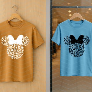 May include: Two short-sleeved t-shirts, one mustard yellow and one light blue, each featuring a Minnie Mouse design. The design includes a leopard print head and bow. The shirts are hanging on wooden hangers.