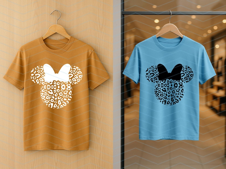 May include: Two short-sleeved t-shirts, one mustard yellow and one light blue, each featuring a Minnie Mouse design. The design includes a leopard print head and bow. The shirts are hanging on wooden hangers.