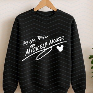 May include: A black long-sleeve sweatshirt featuring a white graphic print that reads Your Pal, Mickey Mouse, accompanied by a small white Mickey Mouse head silhouette. The fabric has a subtle, repeating dark grey chevron pattern throughout.