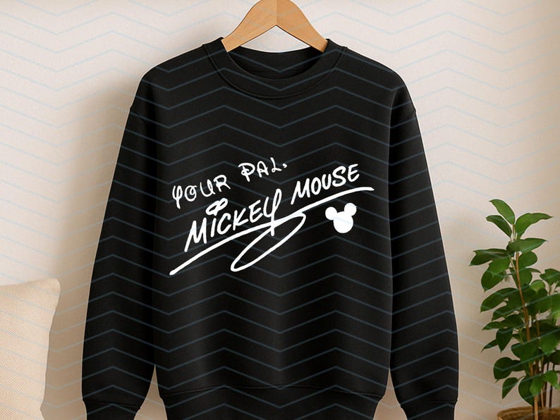 May include: A black long-sleeve sweatshirt featuring a white graphic print that reads Your Pal, Mickey Mouse, accompanied by a small white Mickey Mouse head silhouette. The fabric has a subtle, repeating dark grey chevron pattern throughout.