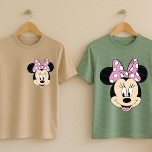 May include: Two t-shirts, one tan and one green, each featuring a Minnie Mouse graphic. The graphic shows Minnie's face with a pink polka-dot bow. The shirts are hanging on wooden hangers.