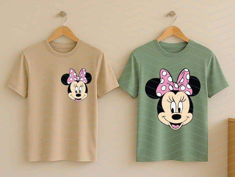 May include: Two t-shirts, one tan and one green, each featuring a Minnie Mouse graphic. The graphic shows Minnie's face with a pink polka-dot bow. The shirts are hanging on wooden hangers.