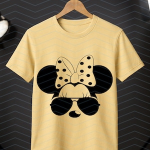 May include: A light yellow t-shirt featuring a black Minnie Mouse design. The design includes sunglasses, a polka-dot bow, and mouse ears. The t-shirt is hanging on a wooden hanger.