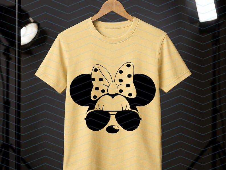 May include: A light yellow t-shirt featuring a black Minnie Mouse design. The design includes sunglasses, a polka-dot bow, and mouse ears. The t-shirt is hanging on a wooden hanger.
