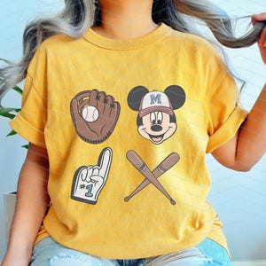May include: A yellow t-shirt with a baseball glove, baseball bat, and Mickey Mouse graphic. The Mickey Mouse graphic has a baseball cap on and says "M" on it. The t-shirt also has a number "1" with a finger pointing up.