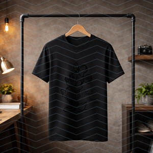 May include: Black t-shirt with a line drawing of a stack of hearts. The shirt is hanging on a wooden hanger from a black metal clothing rack. The background includes a wooden desk, shelves, and a decorative wall.