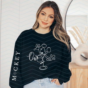 Puede incluir: Una sudadera negra de manga larga con un dibujo de l&iacute;nea continua blanca de Mickey Mouse en la parte delantera y el texto "MICKEY" impreso verticalmente en letras may&uacute;sculas blancas a lo largo del lado exterior de la manga derecha.
